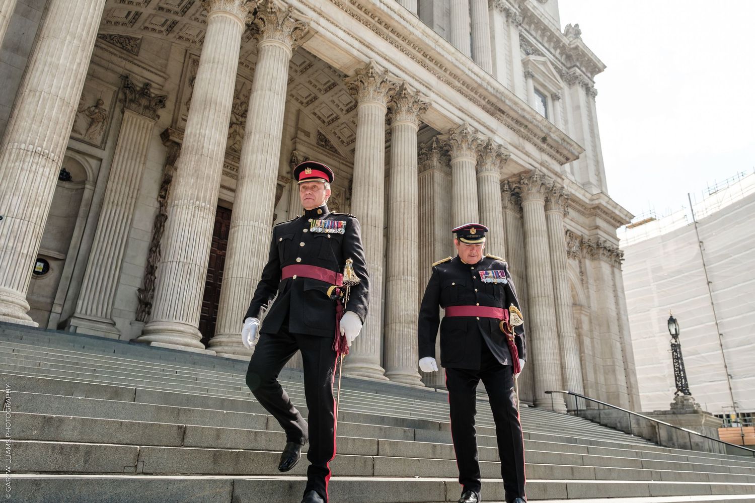 Two military officers in formal uniforms descend grand stone steps outside a neoclassical building with tall columns.
