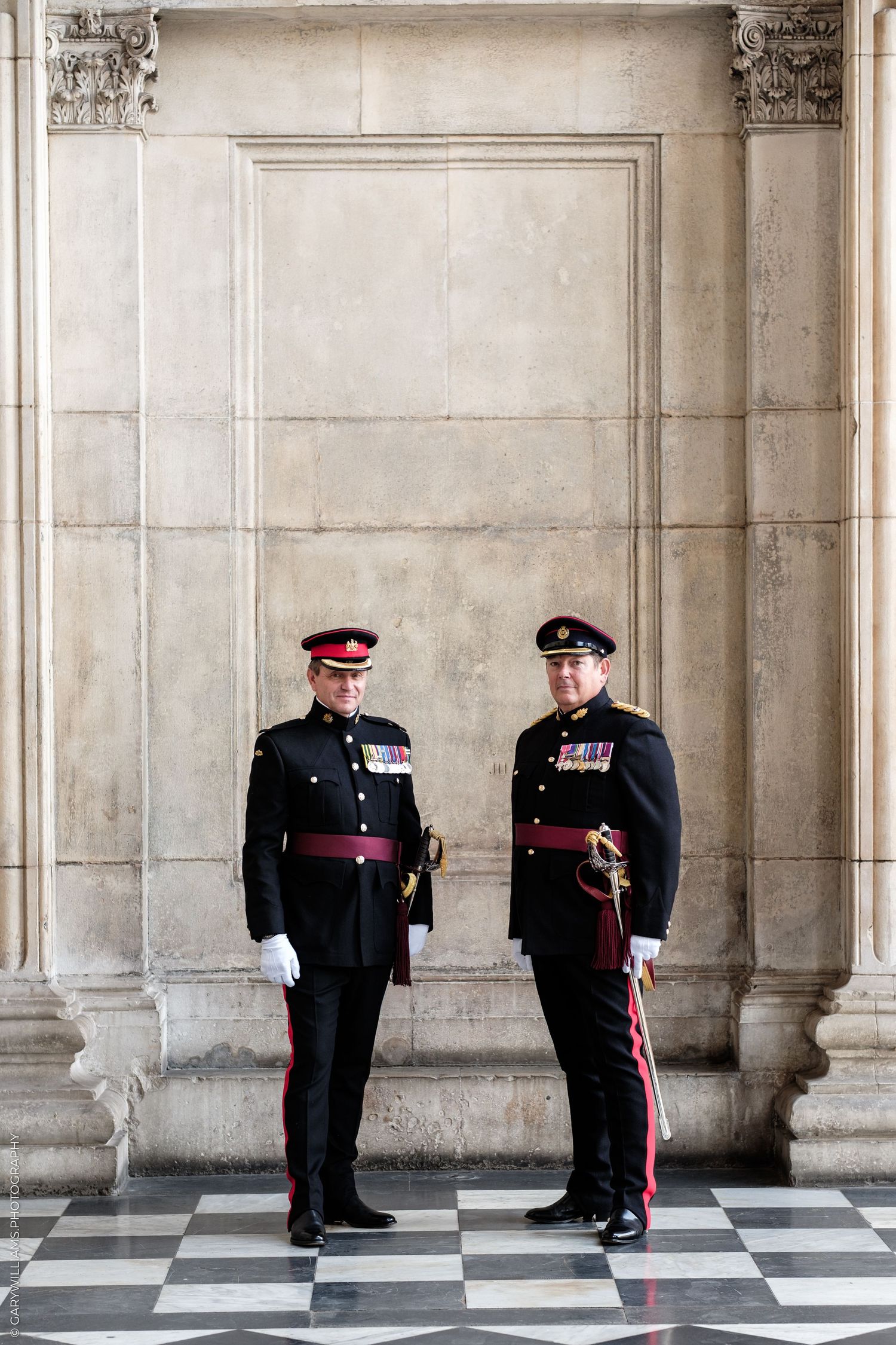 Two military officers in formal black uniforms with red accents and medals stand on a checkered floor before stone columns.