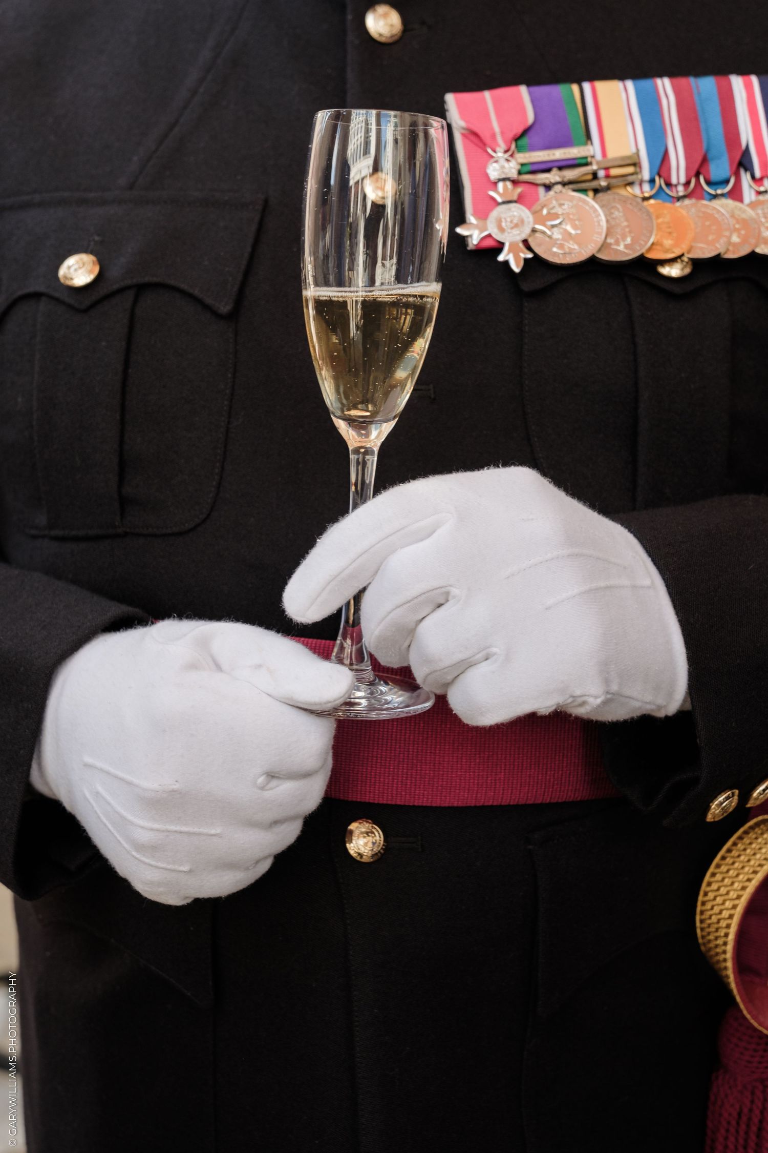 Military officer in dress uniform with medals holding a champagne flute with white gloves.