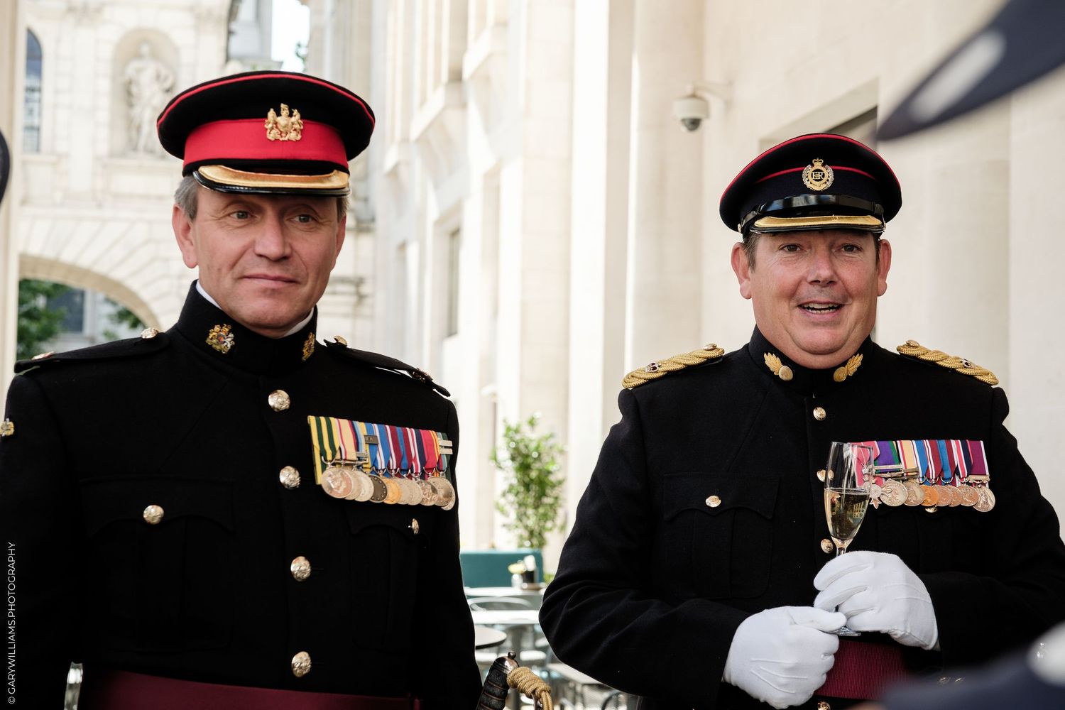 Two men in military dress uniforms with medals smile outdoors at a formal ceremony.