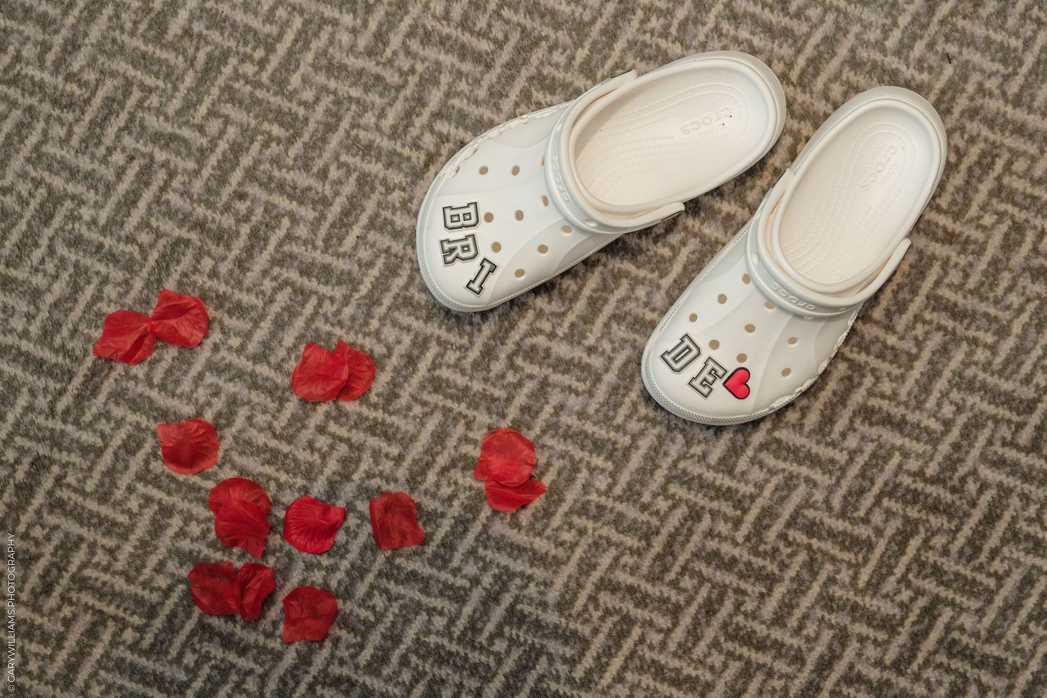 White Crocs shoes with red heart Jibbitz charms on gray carpet with red rose petals arranged nearby.