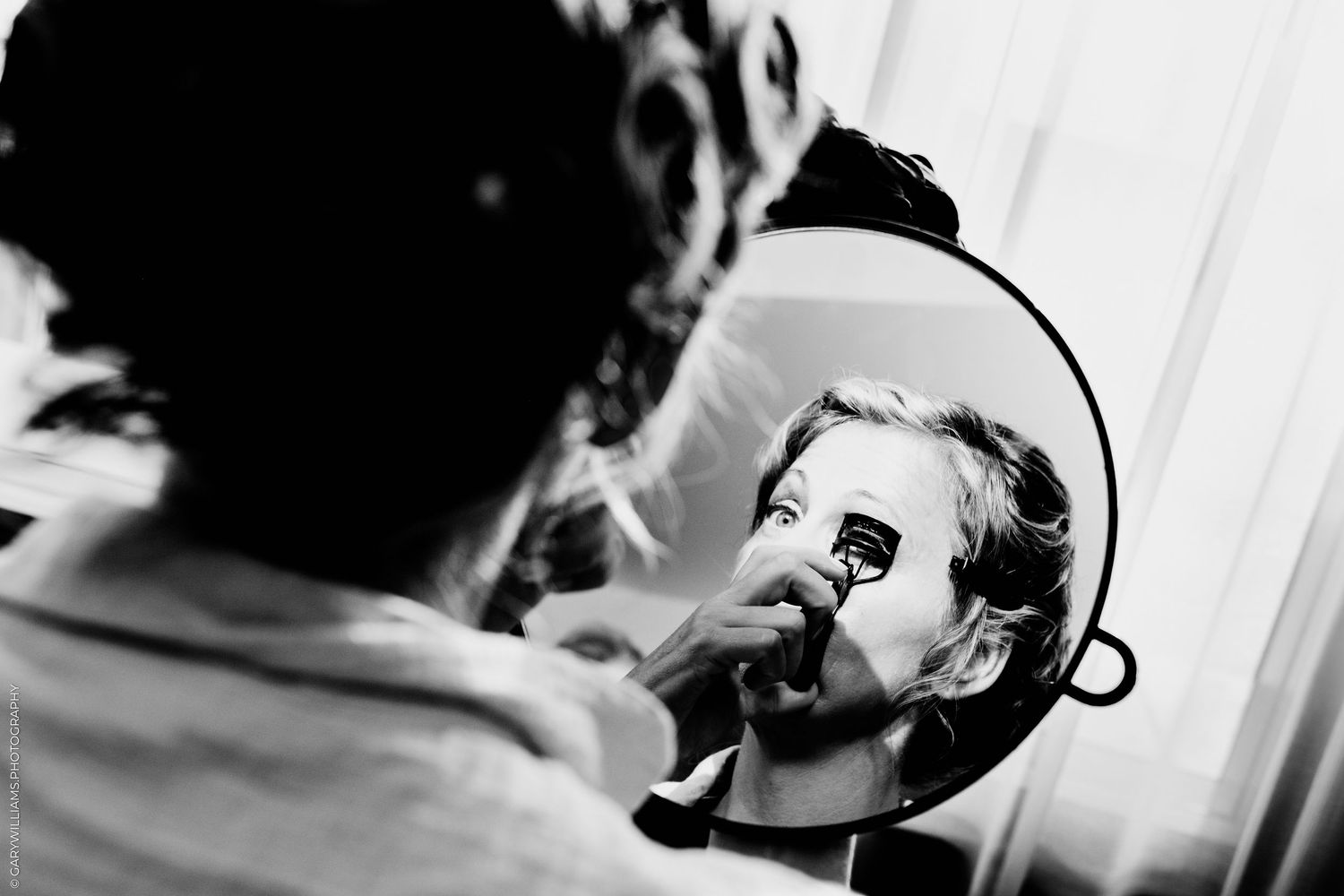 Black and white photo of a woman applying makeup in a round mirror during bridal preparations.