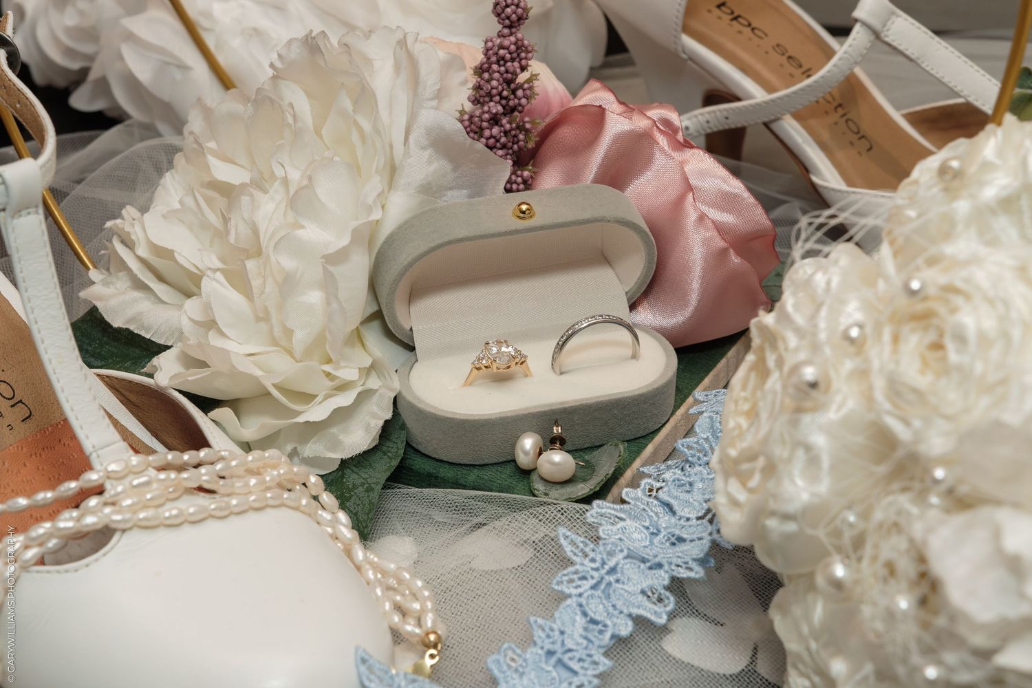 Elegant wedding flat lay with diamond ring in velvet box, pearl necklace, white heels, lace, and floral bouquet.
