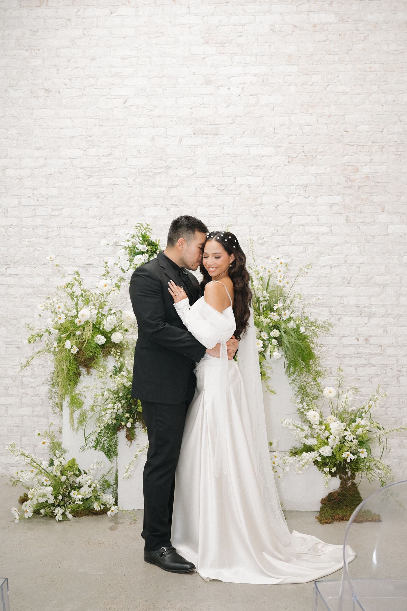 Bride and groom embrace before lush green and white floral arrangements at Oak Atelier in Conroe, Texas.