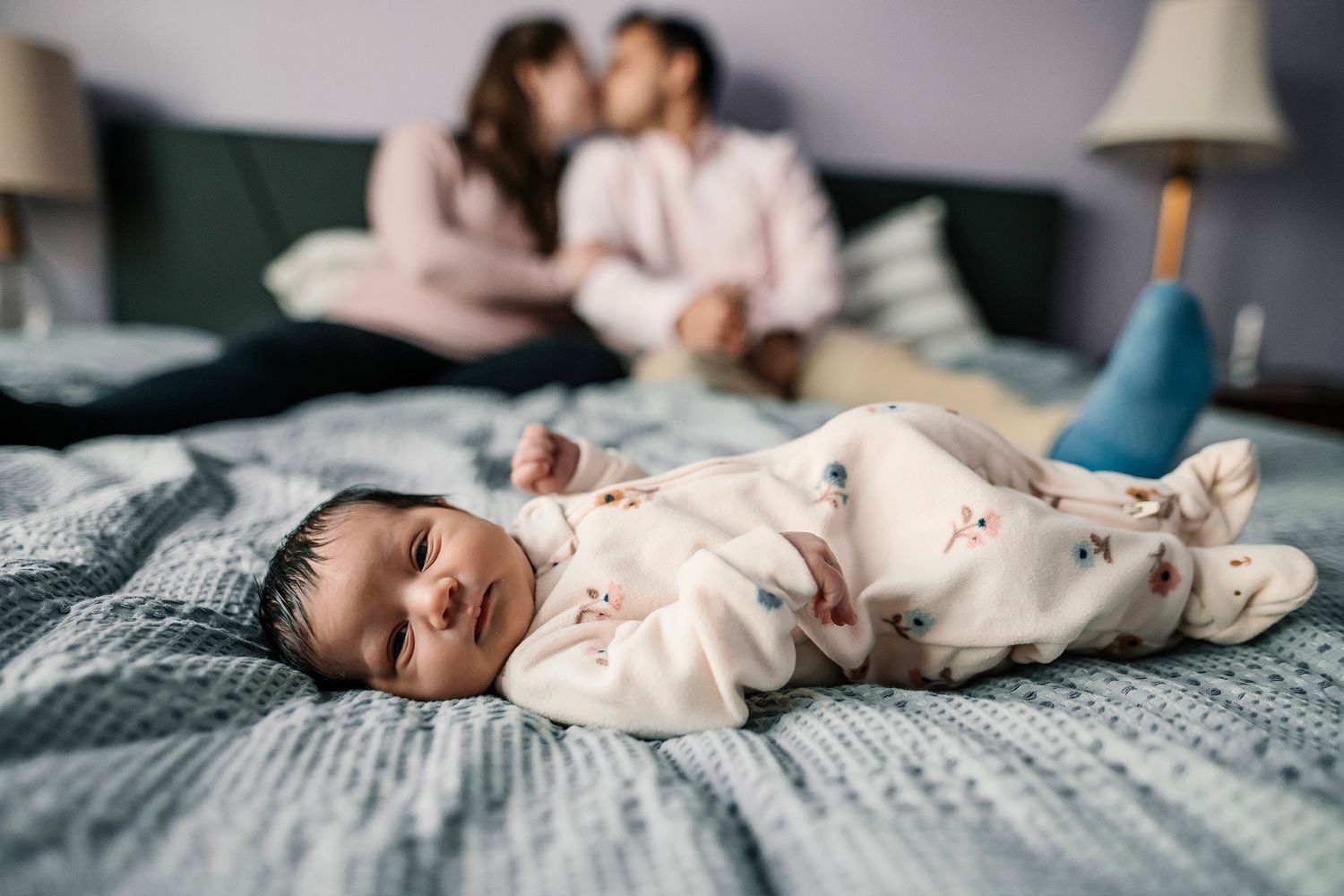 Newborn baby in floral onesie lying on bed with parents kissing softly in the background.