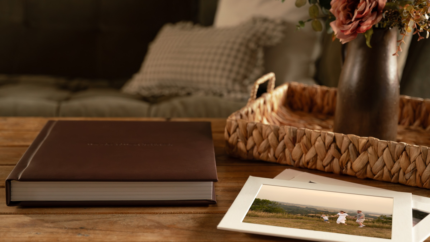 Brown leather photo album and framed landscape print on a rustic wooden table beside a woven tray with dark vase of pink roses.