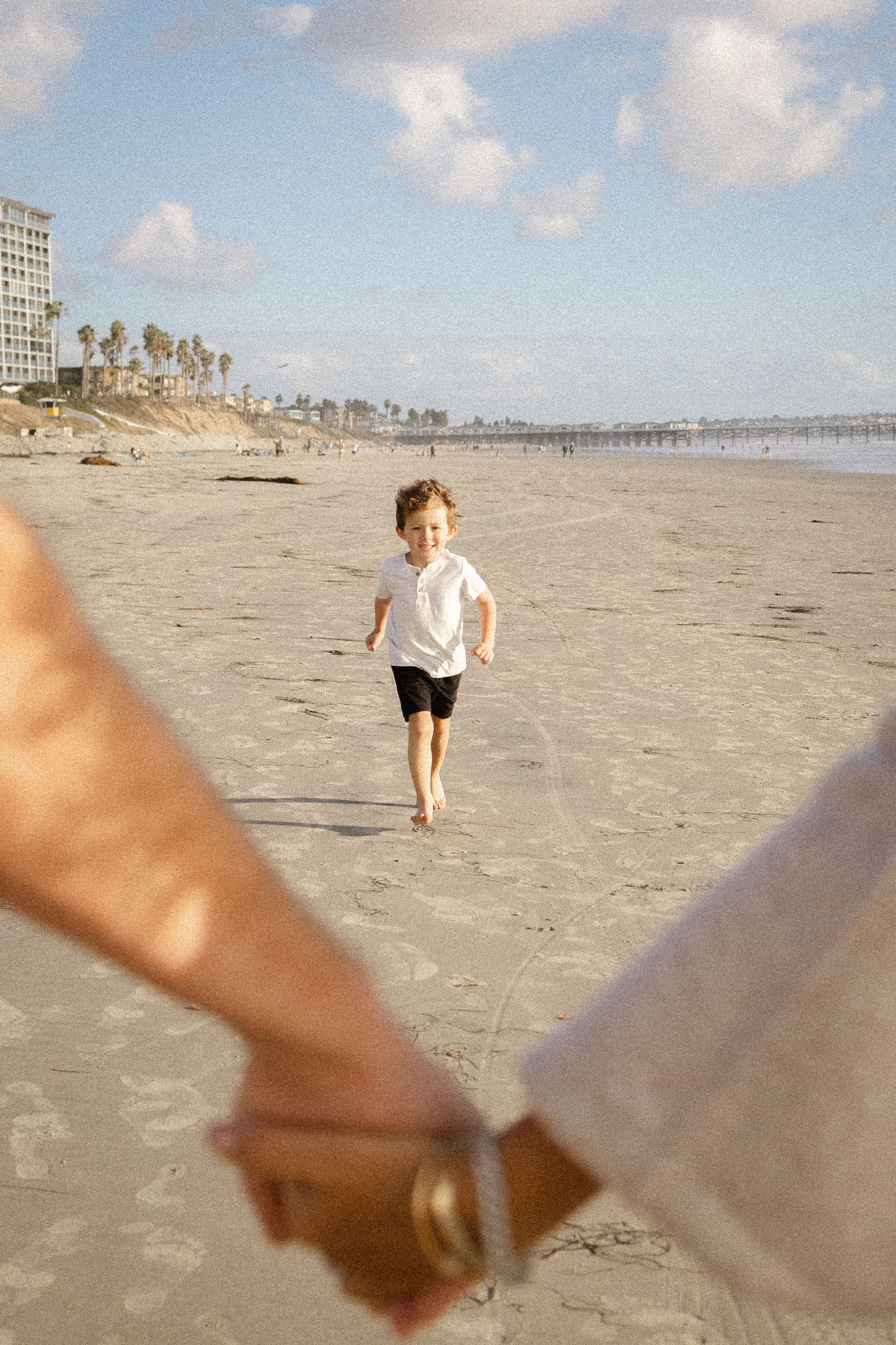 Young boy in a white shirt and black shorts runs toward two people holding hands in the foreground on a sunny sandy beach.