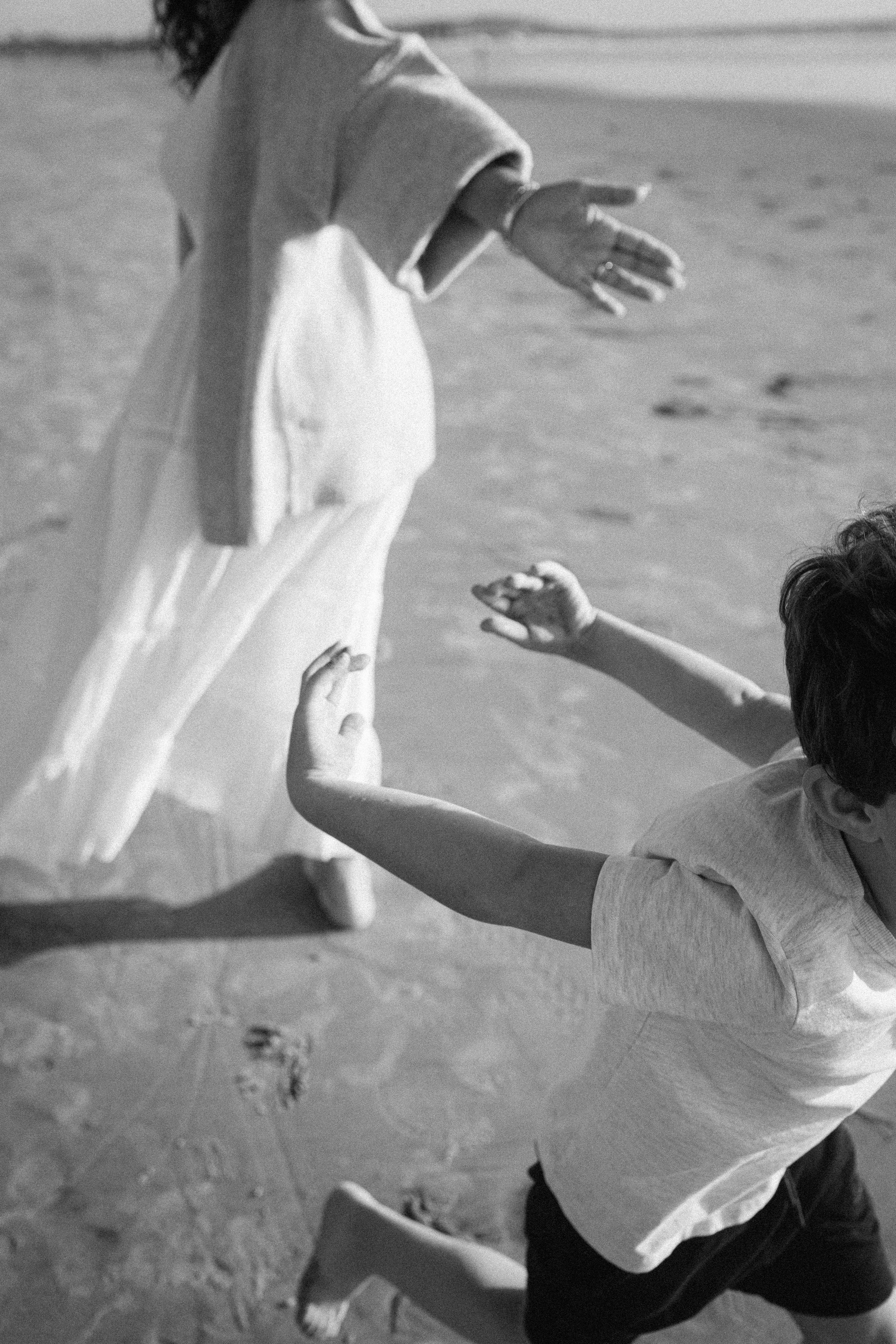 Black and white photo of a woman in a flowing white dress and a child with outstretched arms playing together on a sandy beach.
