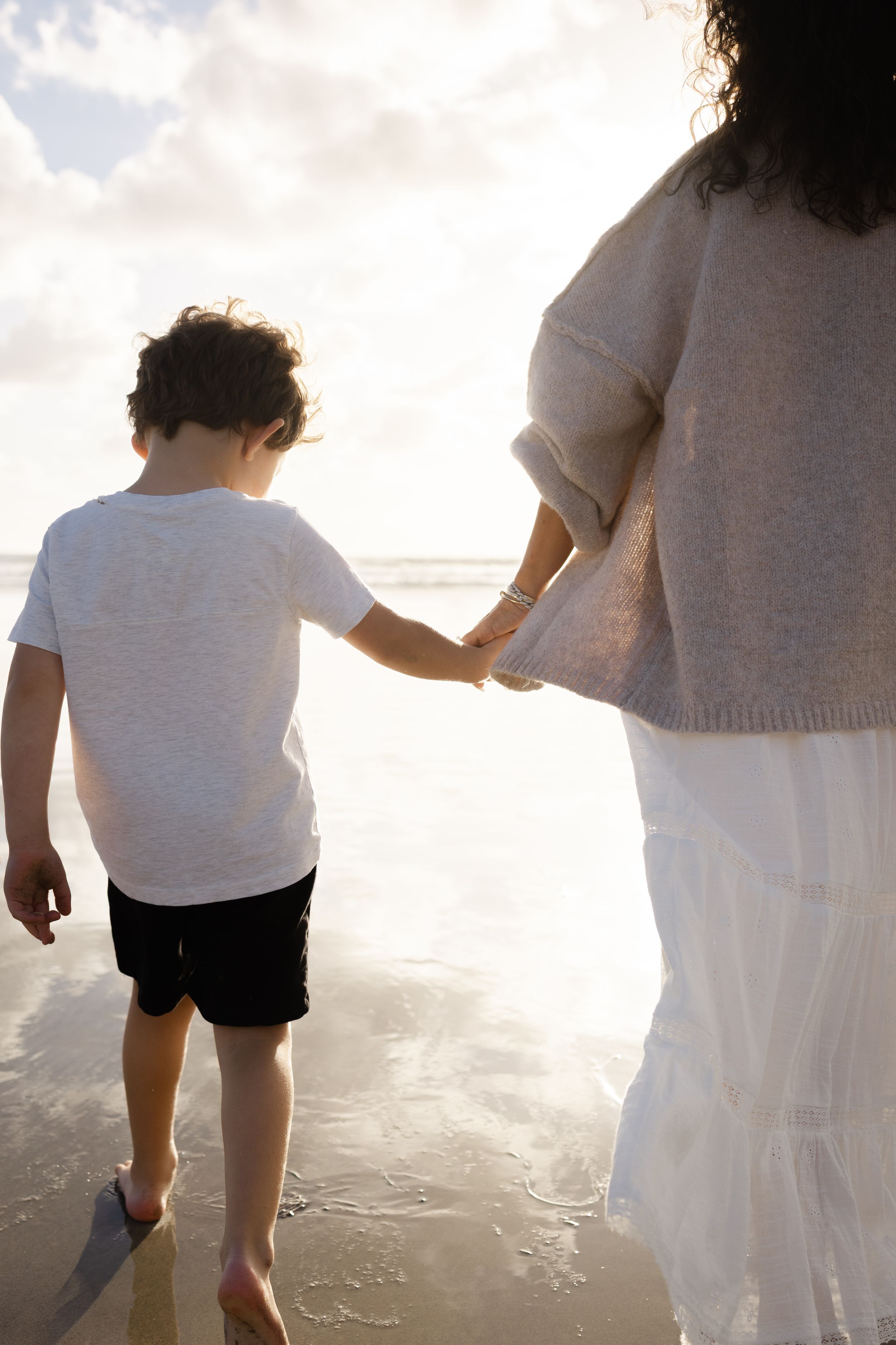 A child in a white t-shirt and black shorts holds hands with a woman in a white skirt while walking barefoot on a sunlit beach.