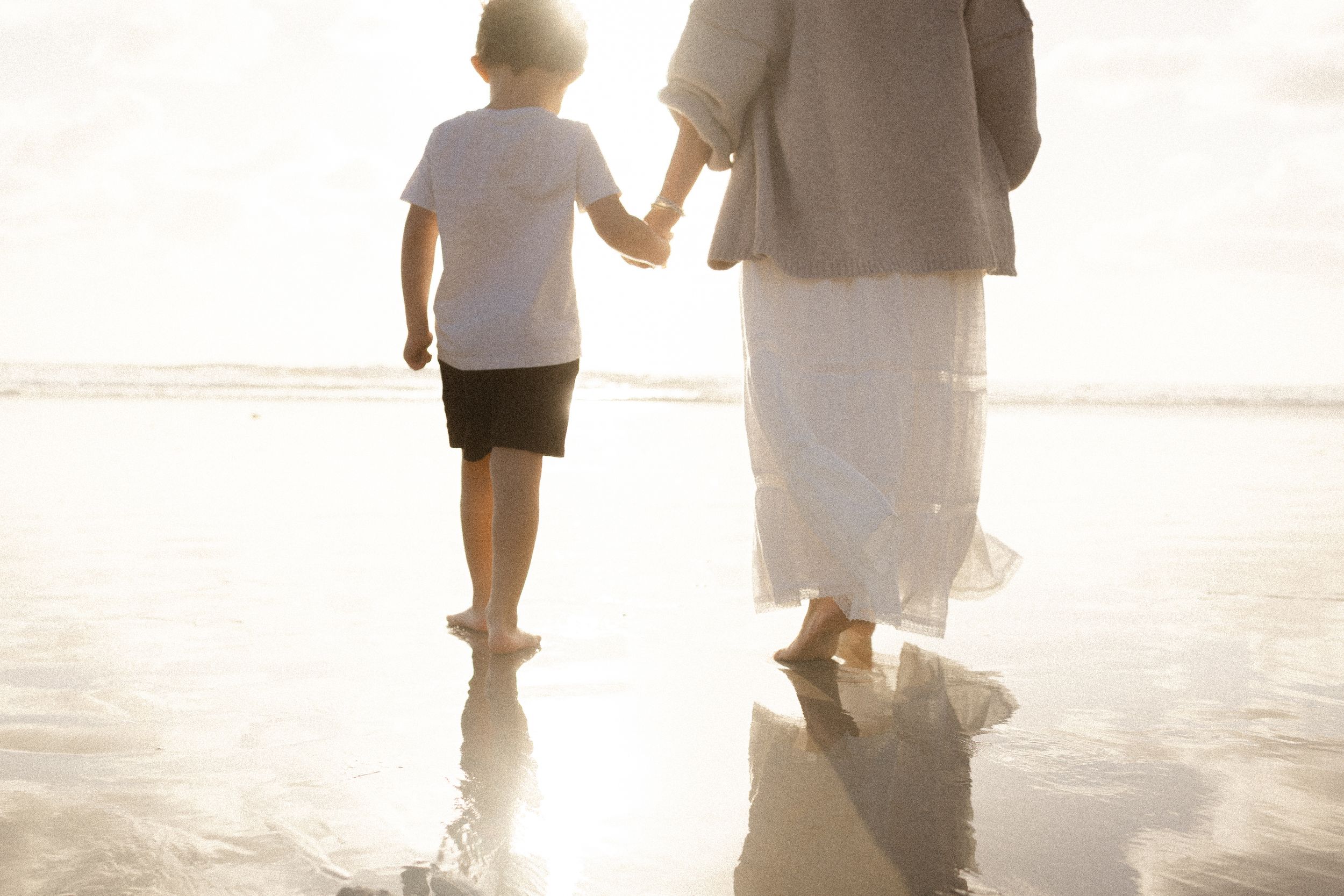Child and adult holding hands while walking barefoot on wet sandy beach at sunset, with golden light reflecting on the shallow water.