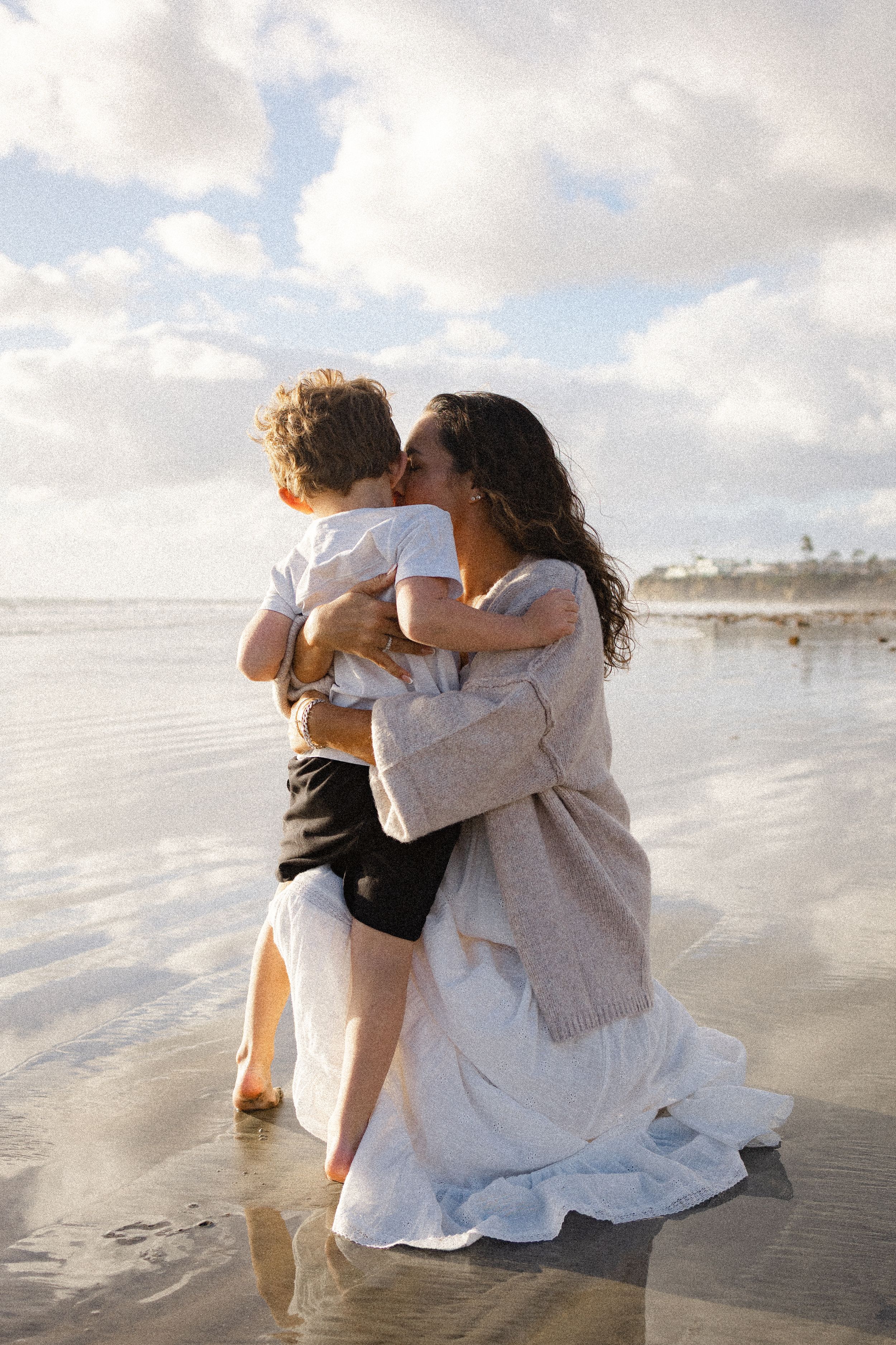 A woman in a white skirt and cardigan kneels on the wet sand and hugs a young boy near the ocean at sunset.