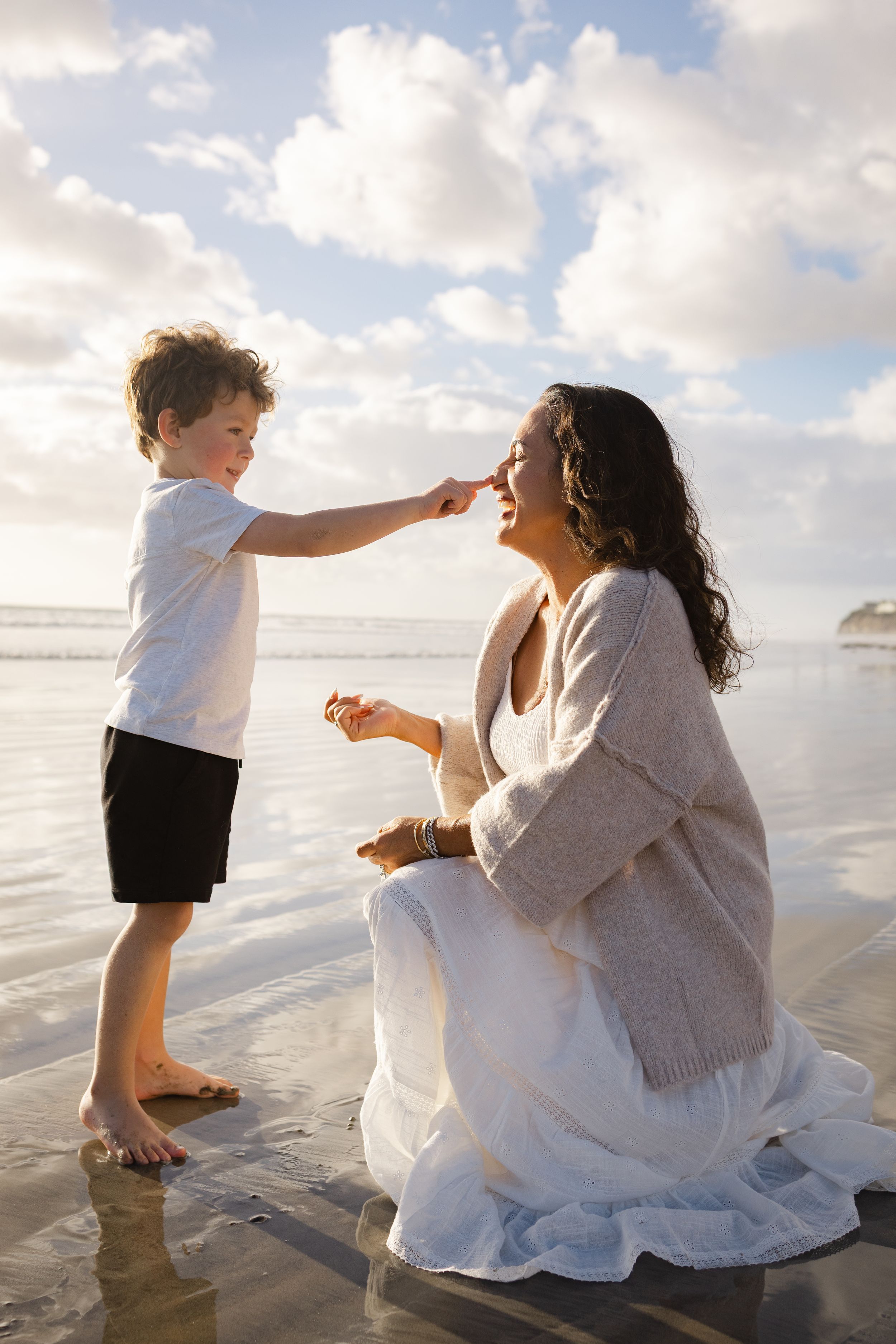 A young boy playfully touches a laughing woman's nose as she kneels in white skirt on a sunlit, reflective beach.
