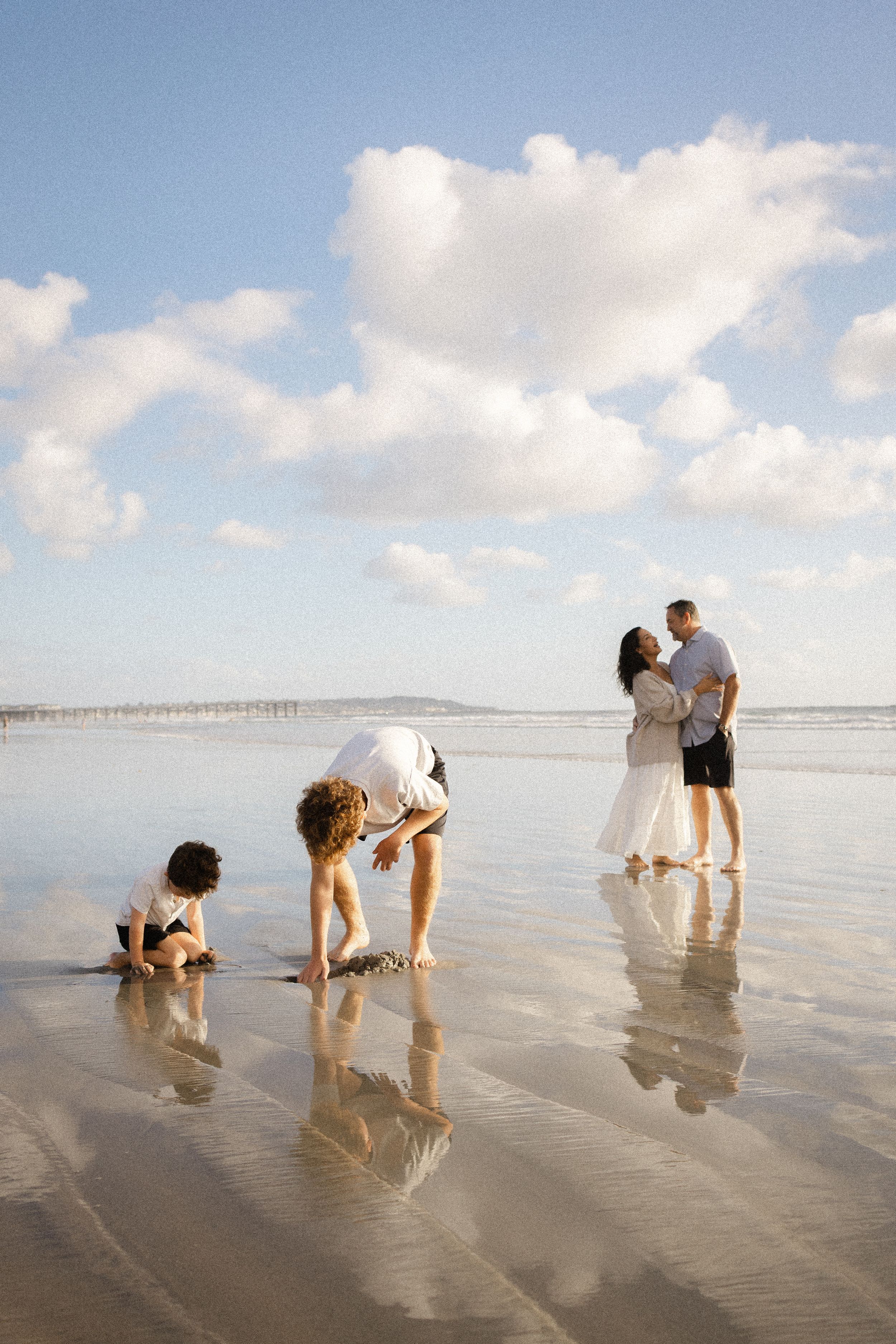 A family plays on a wide, reflective beach under a bright blue sky as a couple embraces and two children dig in the sand.