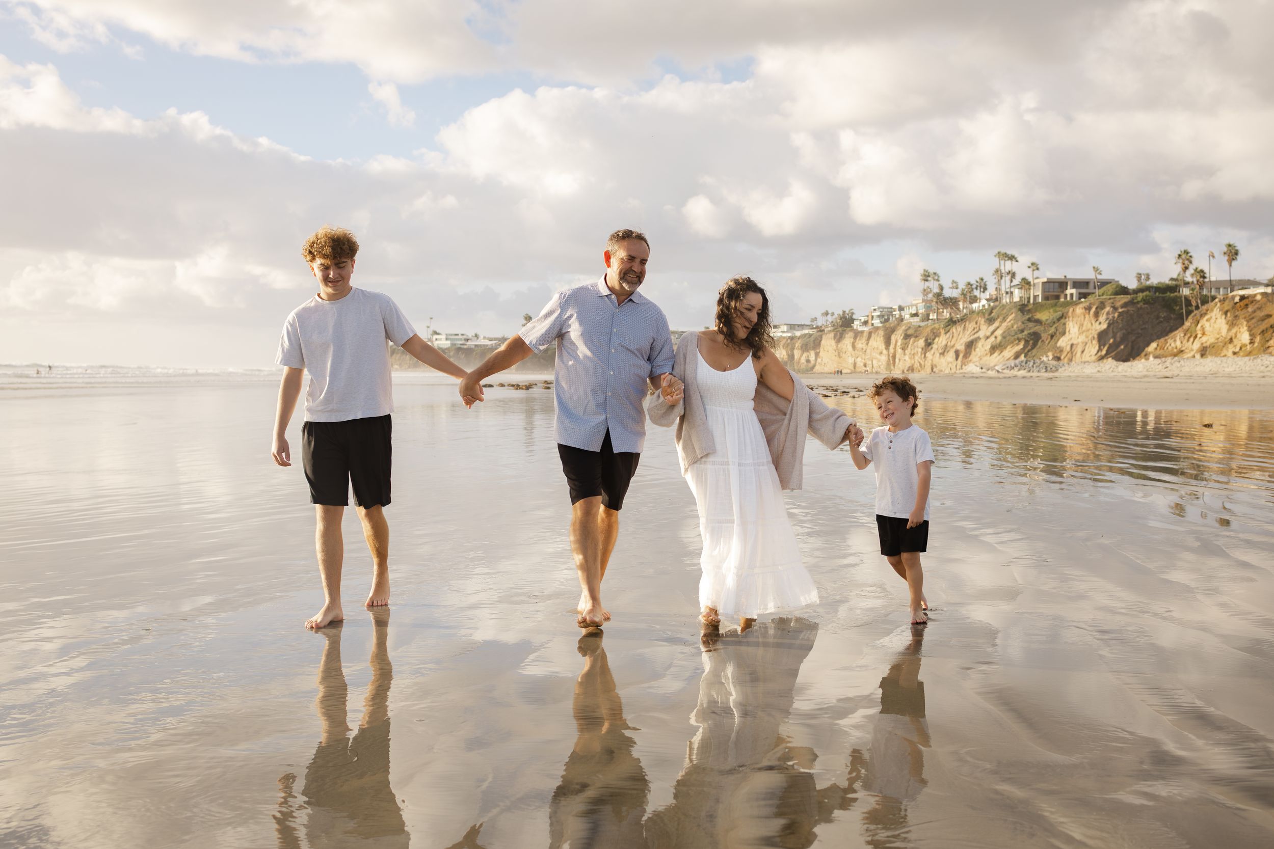 Family of four holding hands and walking barefoot on wet sand at the beach during a golden sunset with cliffs in the background.
