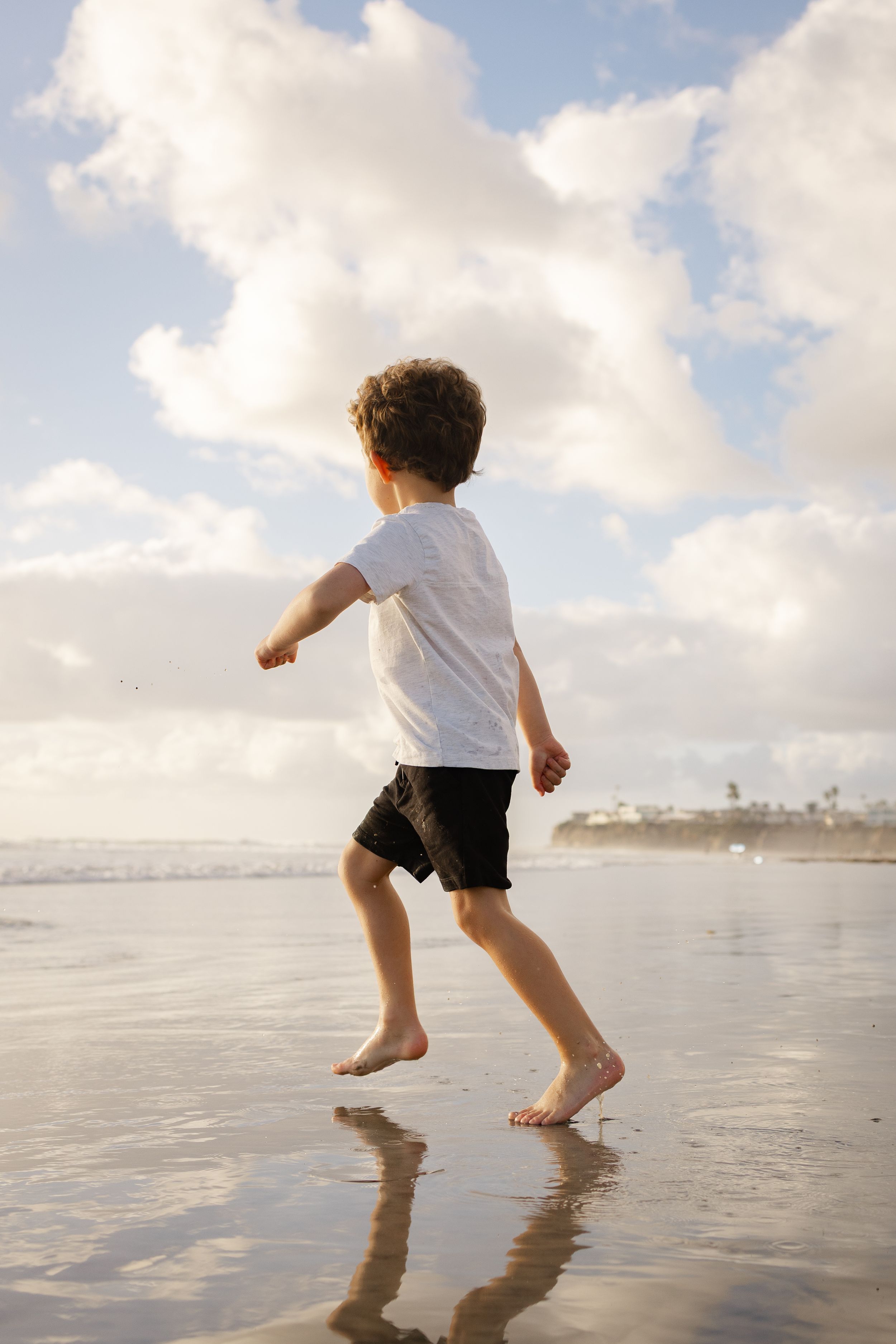 Young boy in a white t-shirt and black shorts jumps barefoot on wet sand at the beach under a cloudy sky at sunset.