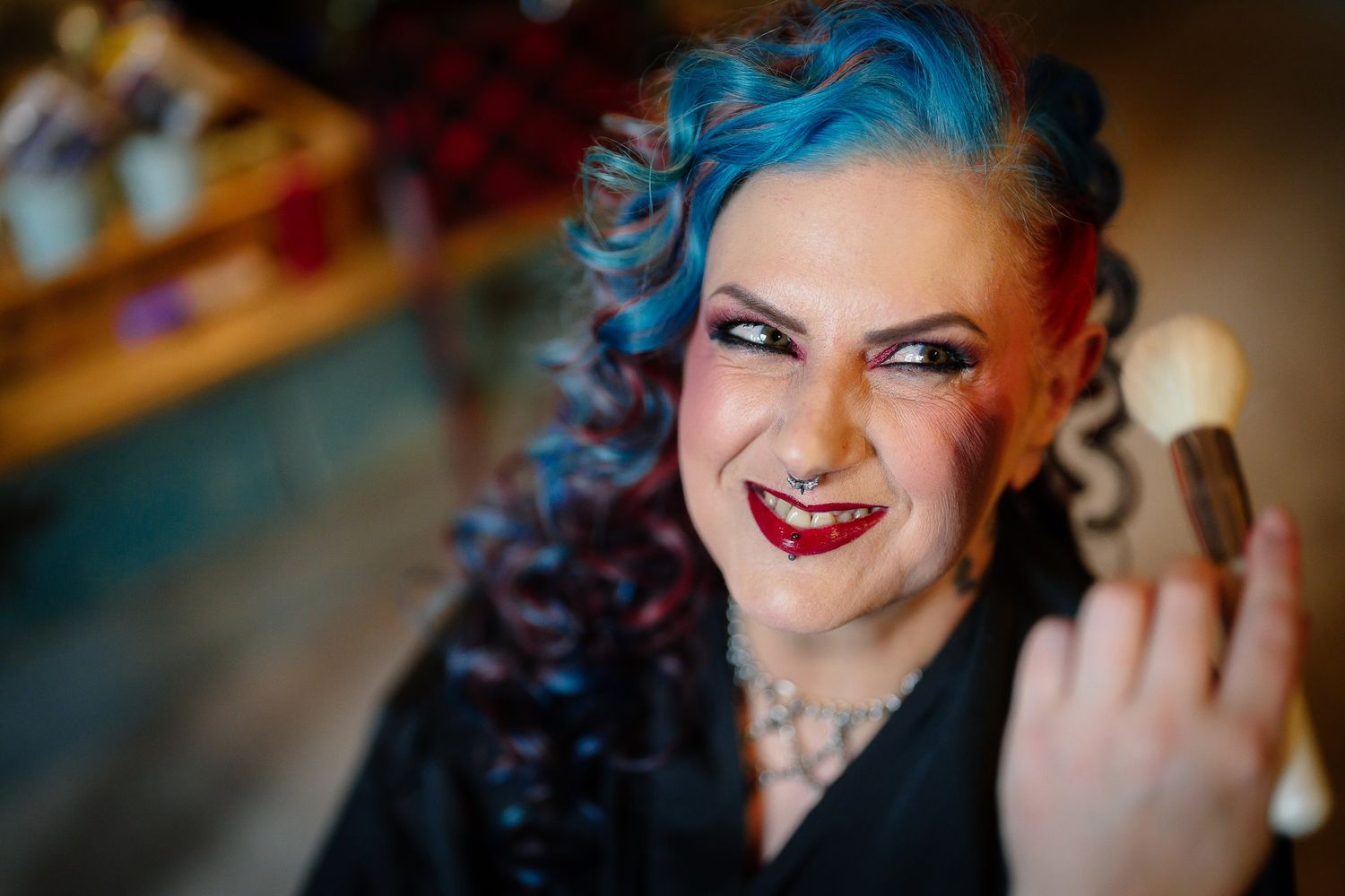 Bride with blue and red curly hair smiling during bridal make-up by Runes and Rouge on her Halloween wedding morning