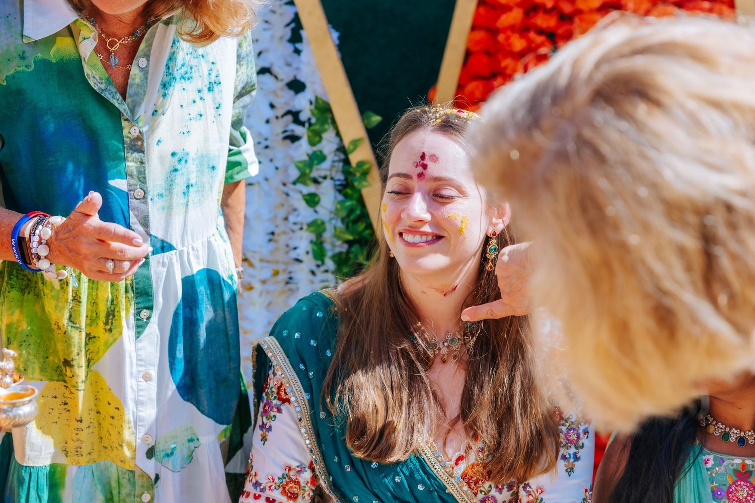 Smiling woman with decorative face gems and floral embroidered teal outfit celebrates at a colorful festive ceremony surrounded by guests.