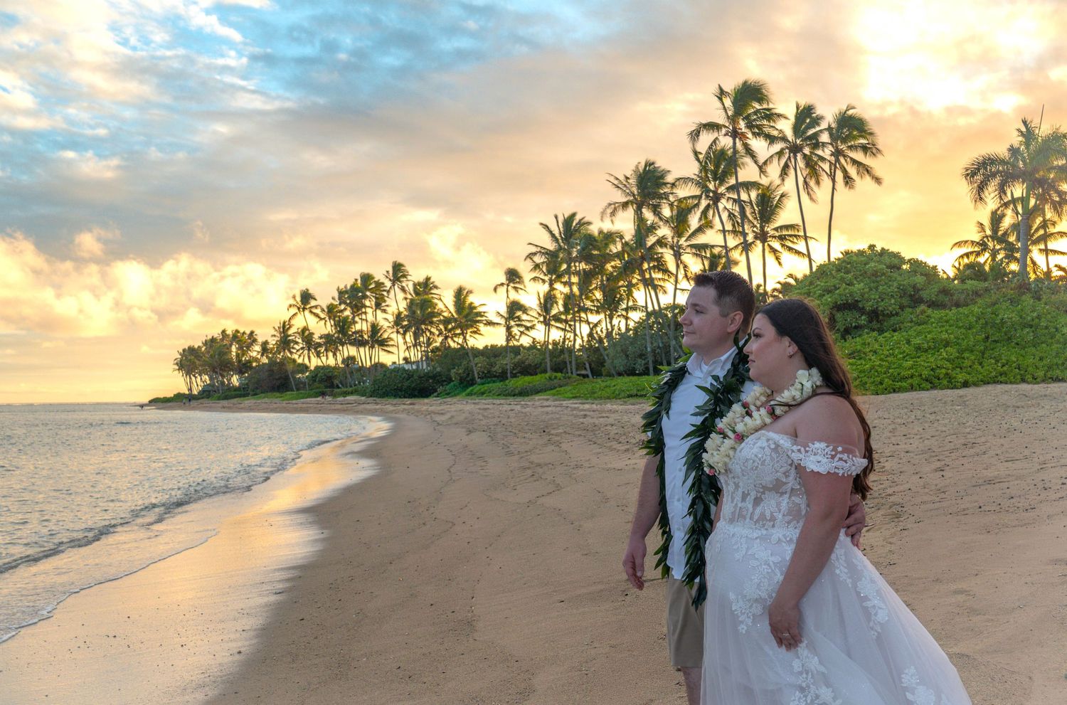 Bride in a white off-the-shoulder gown and groom wearing leis walk along a sandy tropical beach at sunset with palm trees in the background.