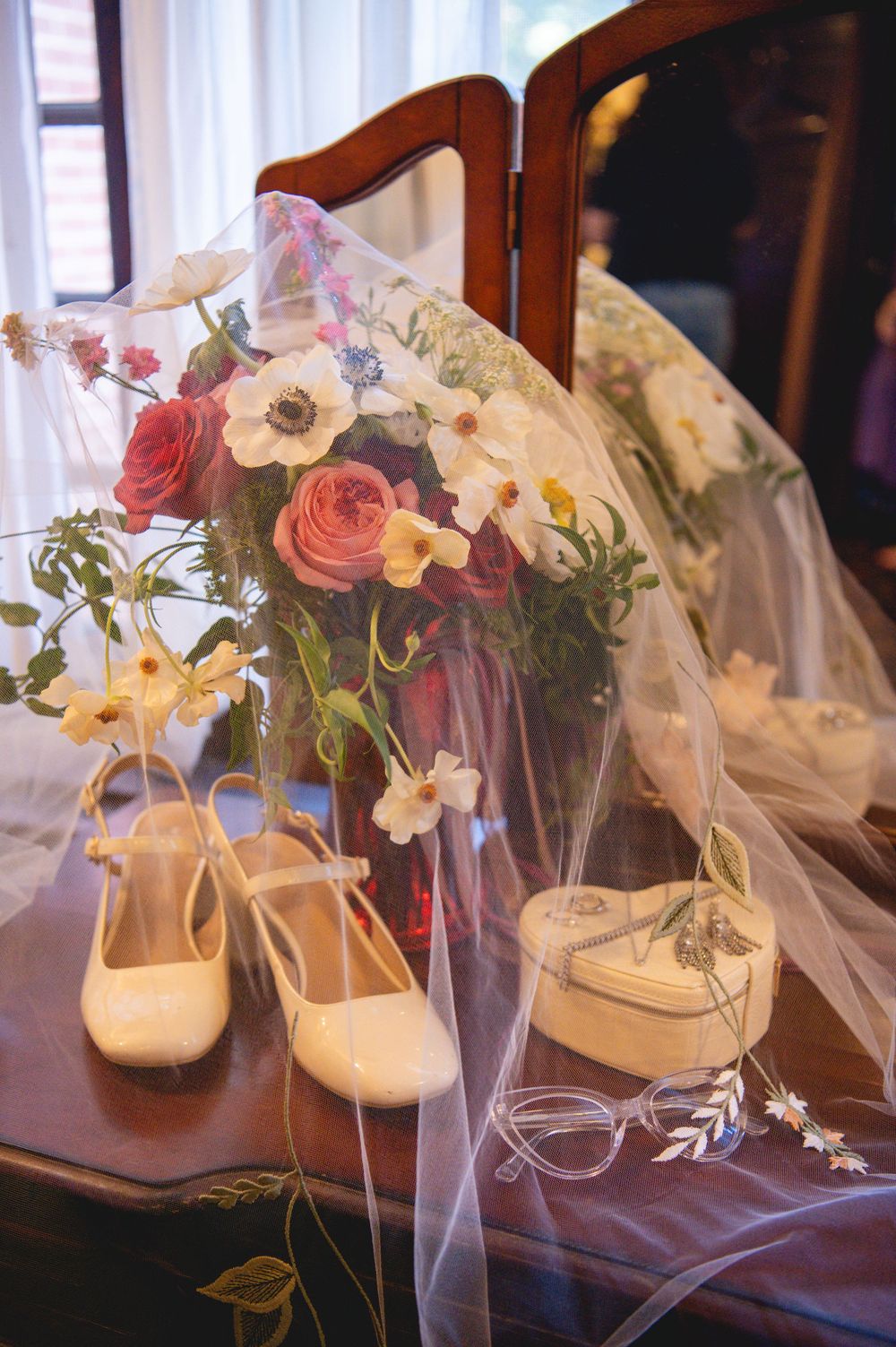 Bridal veil draped over a wooden vanity with cream bridal shoes, a bouquet of roses and anemones, a trinket box, and delicate glasses.