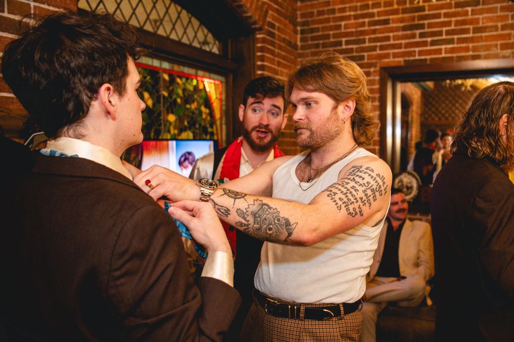 A tattooed man in a white tank top adjusts the collar of another man in a brown jacket while a bearded man smiles behind them.