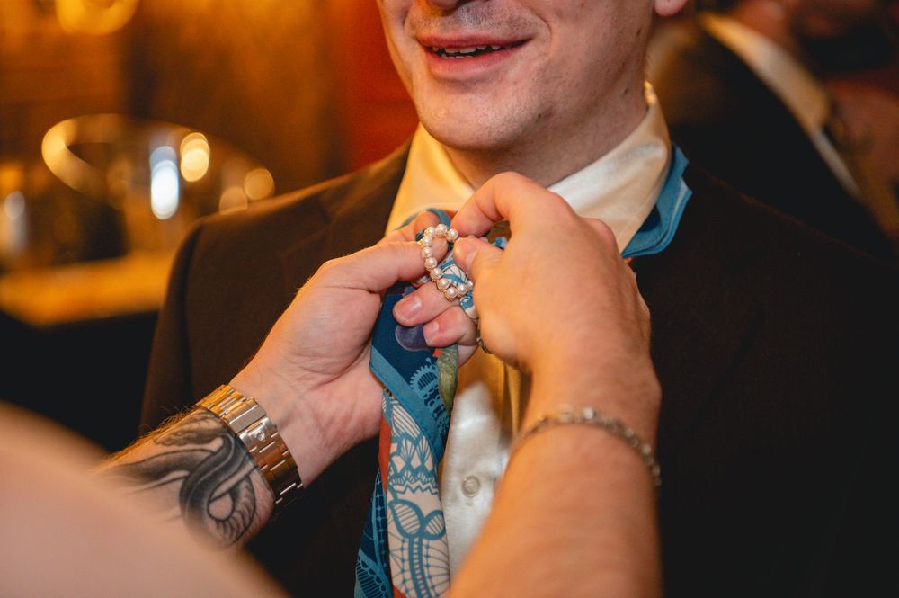 Tattooed hands adjust the patterned blue necktie of a smiling man in a dark blazer and cream shirt at a warm, softly lit indoor event.