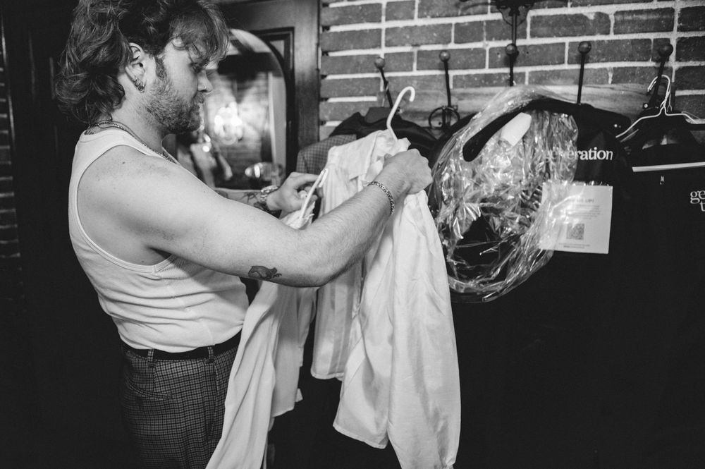 Black and white photo of a bearded man in a tank top sorting through white garments on hangers in a backstage wardrobe area.