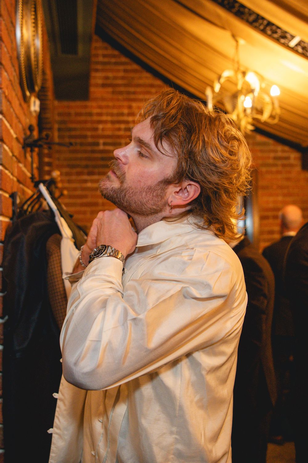 A bearded man with wavy auburn hair adjusts the collar of his cream button-up shirt in a warm brick-walled room with a chandelier.