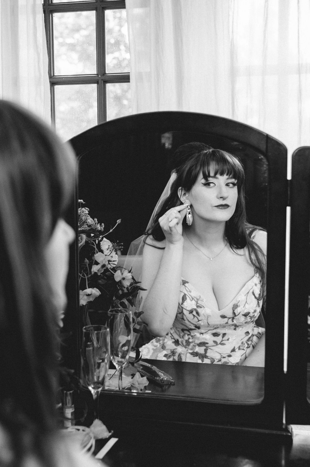 Black and white photo of a woman with bangs in a floral dress putting on an earring while looking into a vanity mirror beside fresh flowers.