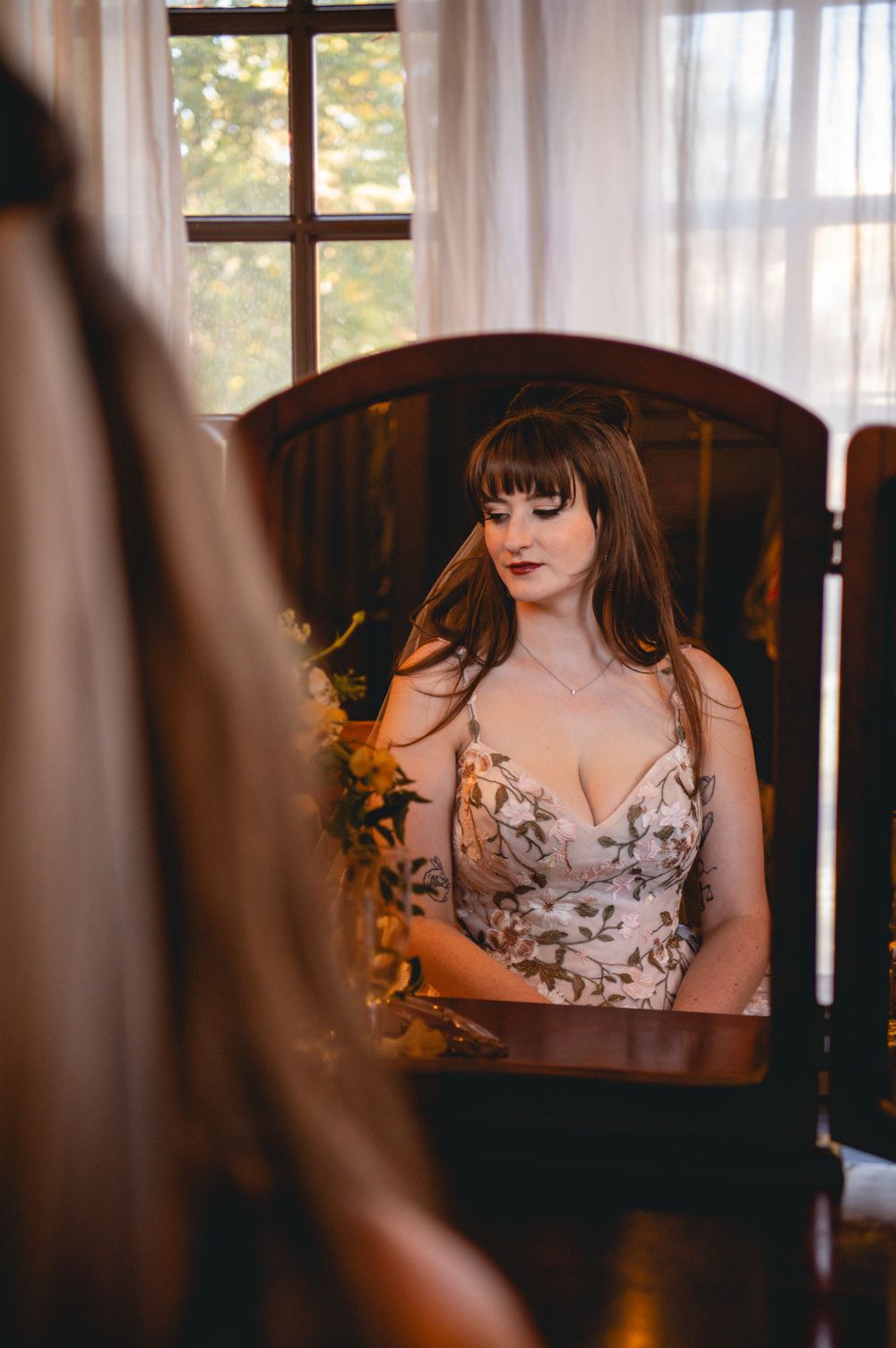Woman with long brown hair and bangs gazes down in a wooden vanity mirror, wearing a floral satin dress beside soft window light.