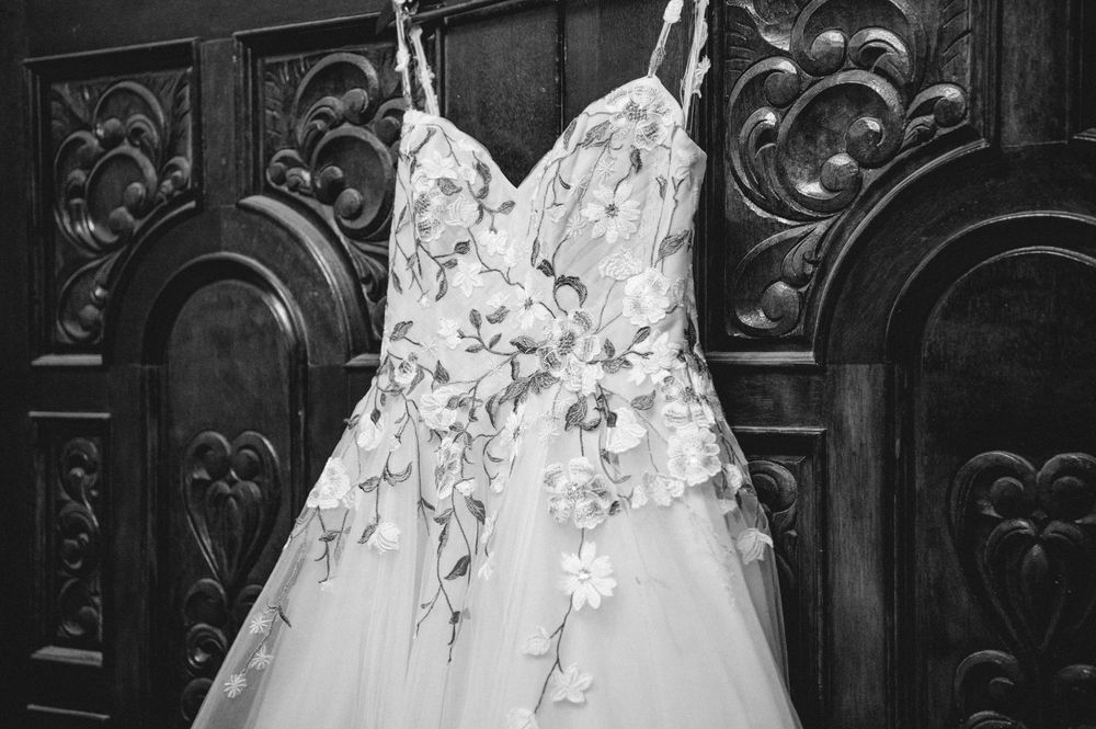 Black and white photo of a floral lace wedding dress with spaghetti straps hanging against an ornate carved wooden door.
