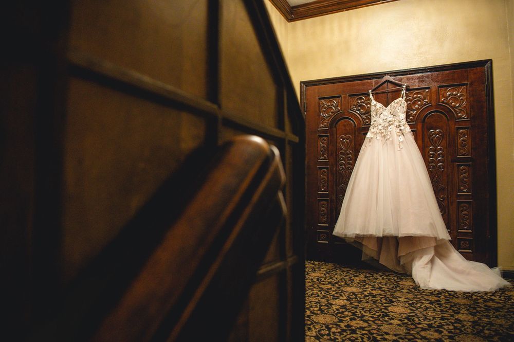 White strapless lace wedding gown with a long train hanging on an ornate carved wooden door beside a dark wooden staircase railing.