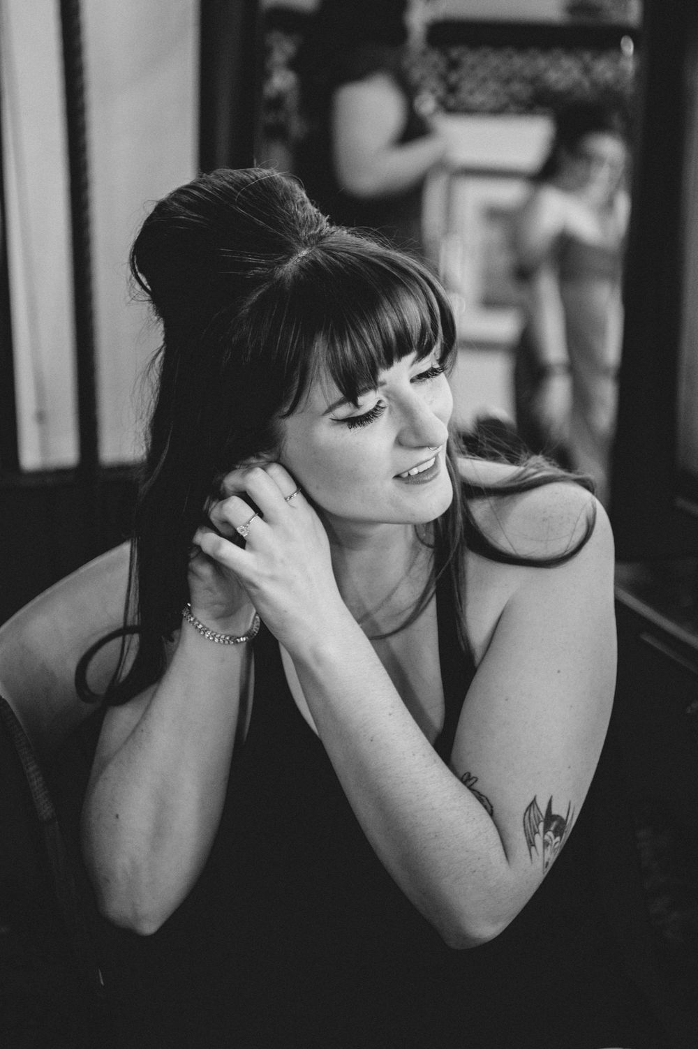Black and white portrait of a smiling woman with bangs and a high updo, touching her hair, with a tattoo on her arm and a mirror behind her.