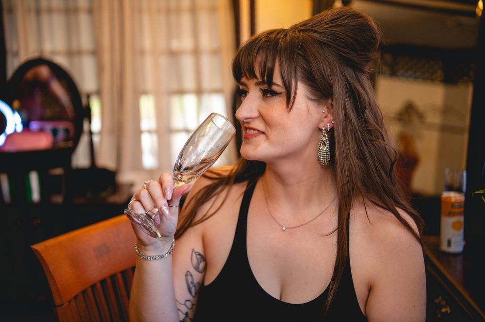 Smiling woman with an updo and tattooed arm holds a champagne flute while seated in a cozy, warmly lit room.