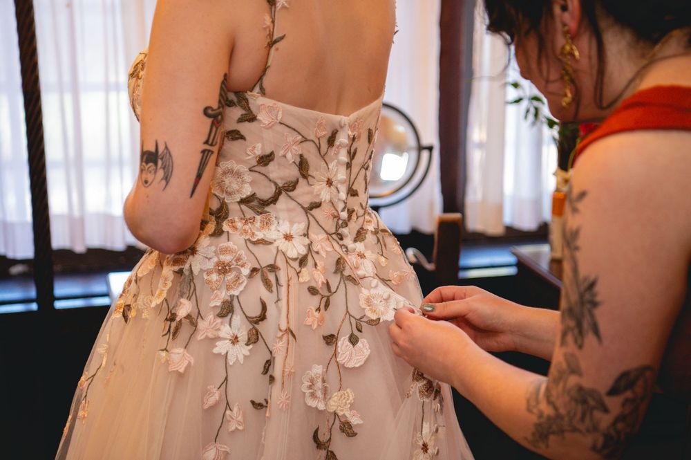 Bride in a blush wedding gown with embroidered floral appliqué has her dress fastened by a tattooed helper in a bright, window-lit room.
