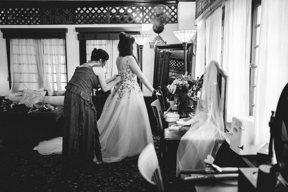 Black and white photo of a bride in a lace wedding gown being helped by a woman as they prepare in a softly lit room.