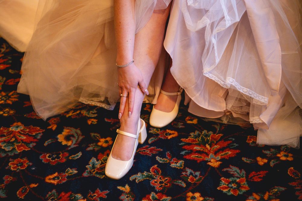 Bride in a white lace gown adjusts her cream Mary Jane wedding shoe while seated on a floral patterned carpet.