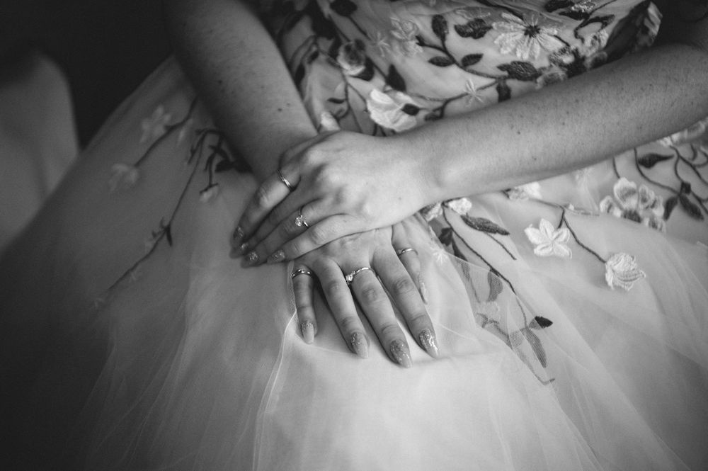 Black and white close-up of a bride's hands with rings resting on her floral embroidered tulle wedding dress.