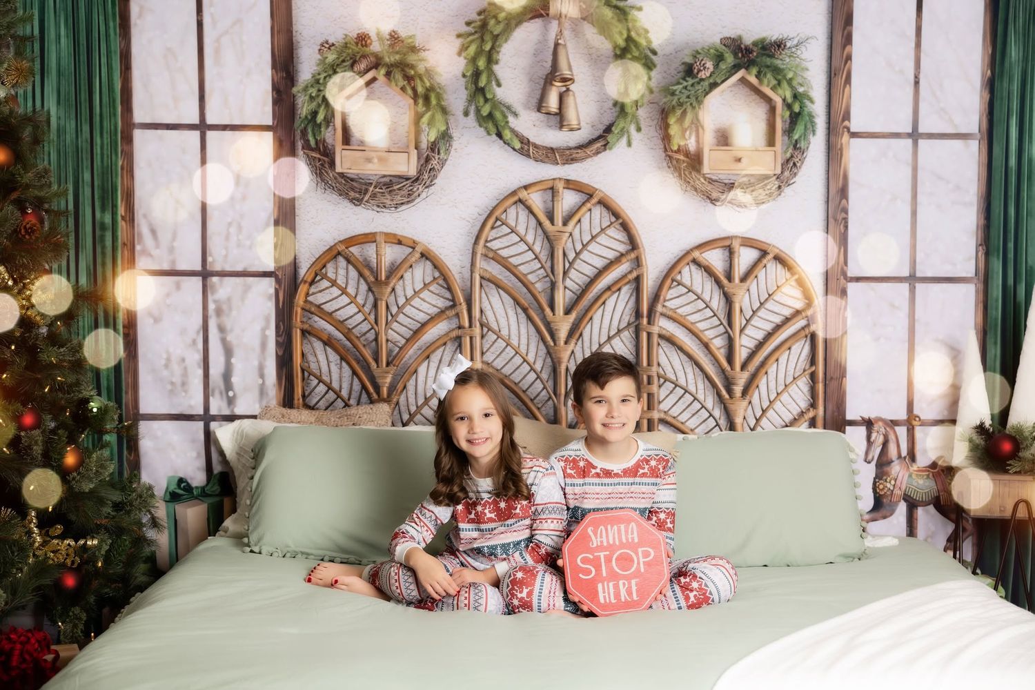 Two children in matching Christmas pajamas sit on a green bed holding a stop sign, with wreaths, wooden headboard and a decorated tree.