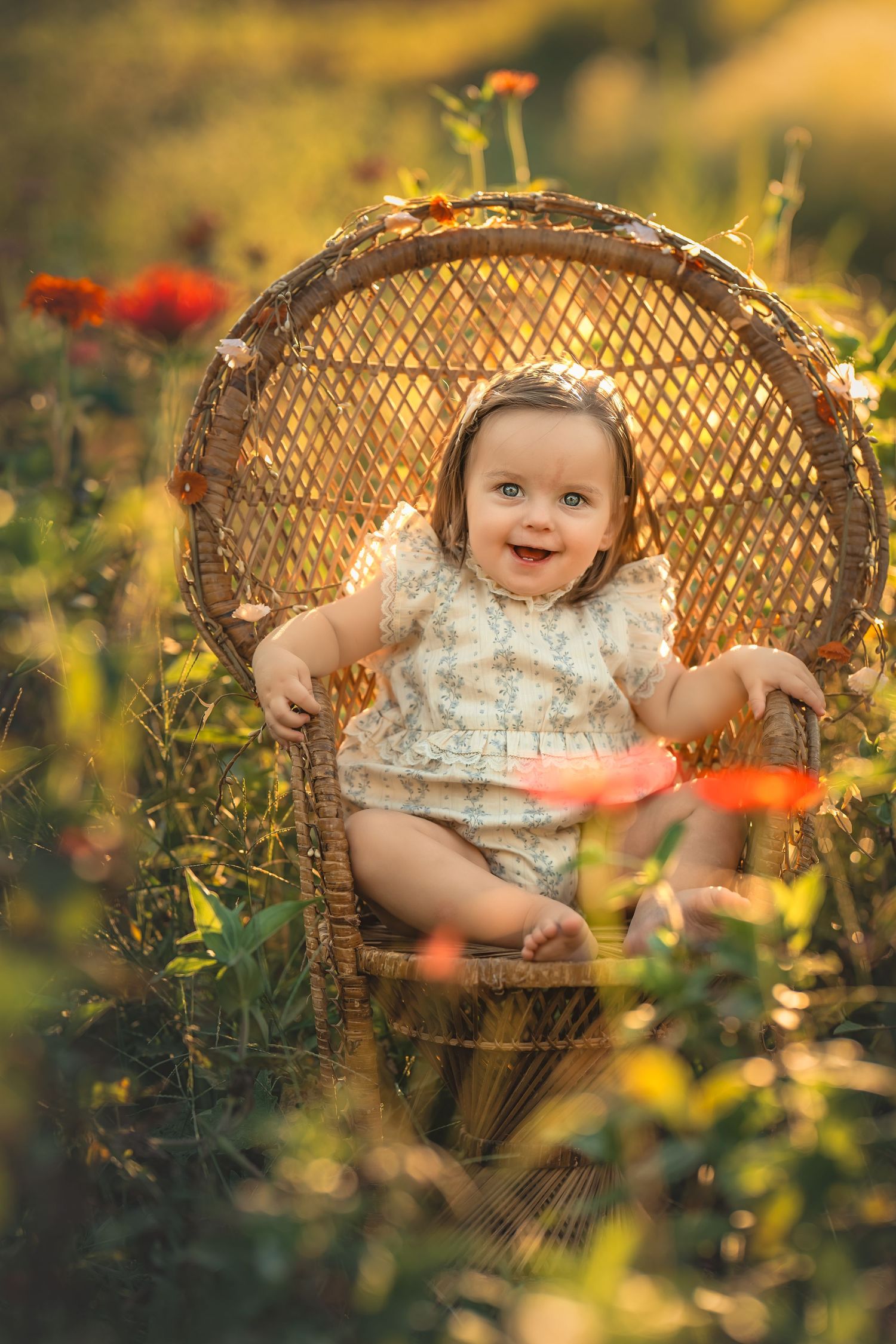 A smiling baby girl sits in a wicker peacock chair surrounded by colorful wildflowers in golden sunlight.