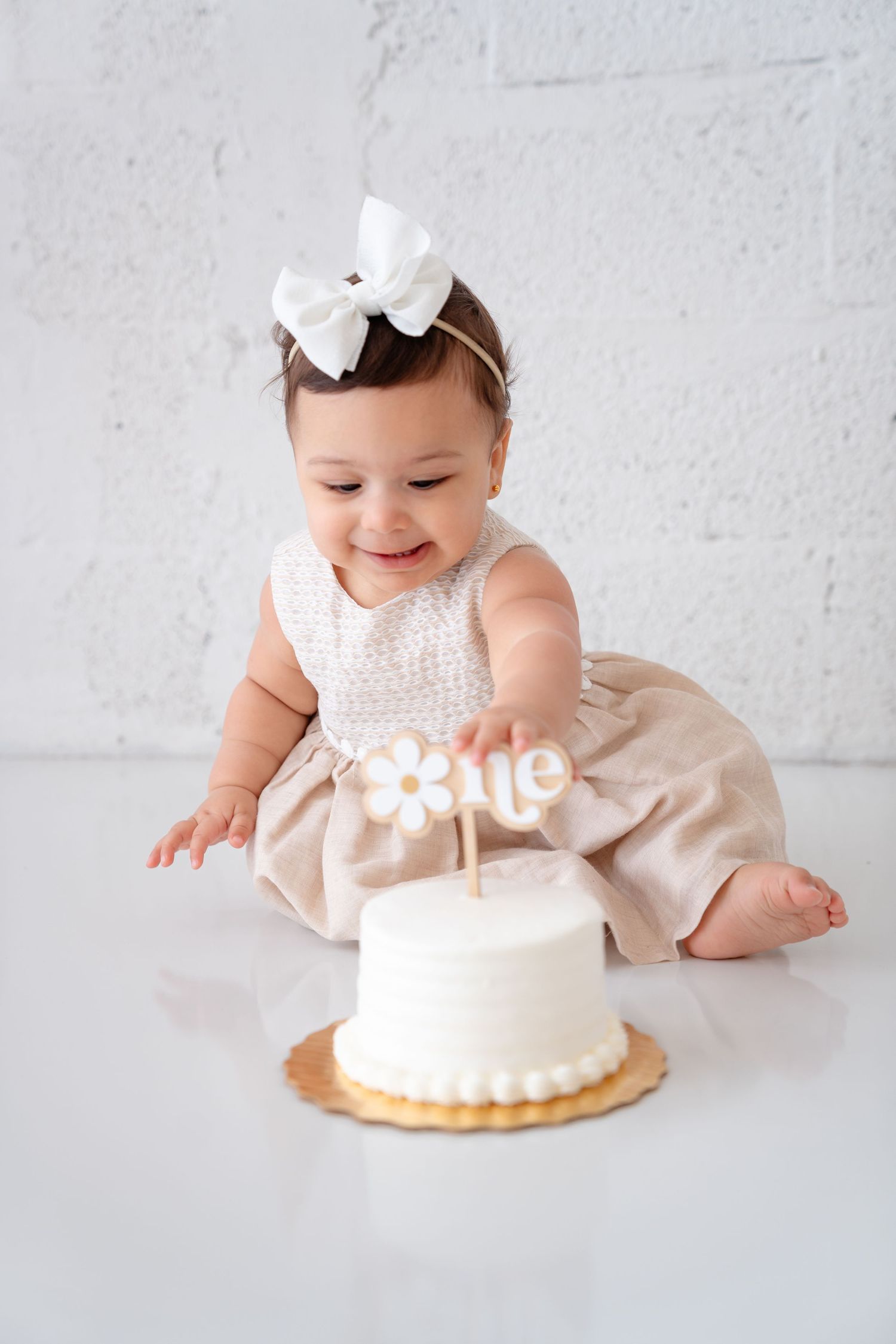 Smiling baby girl in beige dress with white bow celebrates first birthday with white cake topper reading 'one'.
