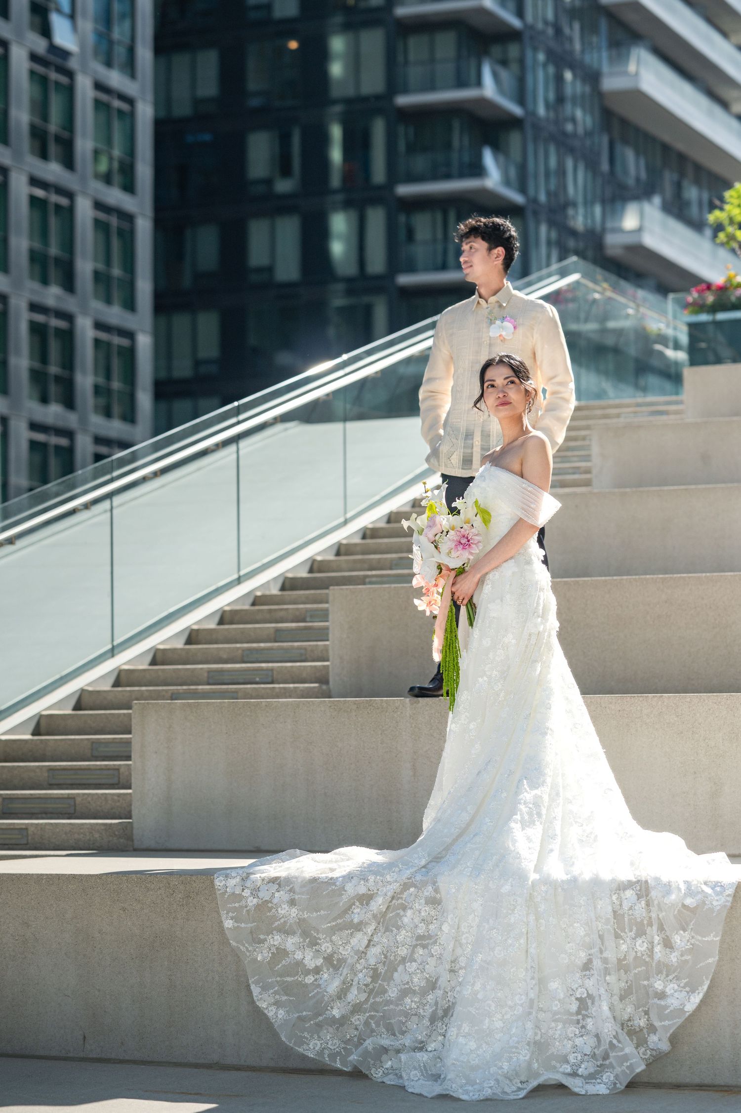Bride in lace gown holding bouquet with groom in beige suit posing on outdoor steps near modern building.