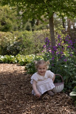 Young girl in white dress smiling while kneeling in a garden with purple flowers and a wicker basket during a mother and daughter session