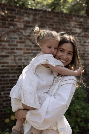 A smiling woman in a white dress holds a young blonde girl in a matching white dress outdoors near a brick wall in Brighton