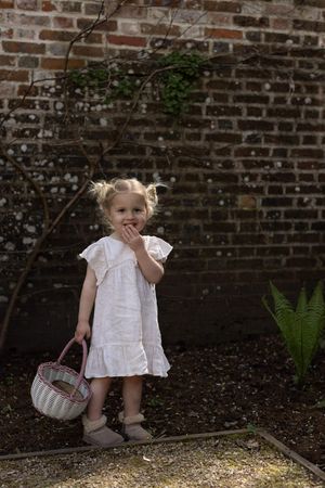 Young girl in white dress holding a pink basket stands in a garden against a rustic brick wall in Brighton