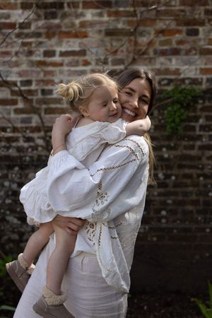 Woman in white embroidered dress smiling while holding a toddler girl in white dress against brick wall in Brighton