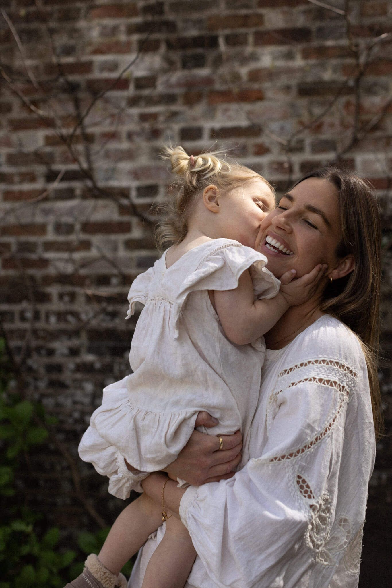 Editorial style photo of toddler girl in white dress kissing her laughing mother on the cheek in white lace dress outdoors