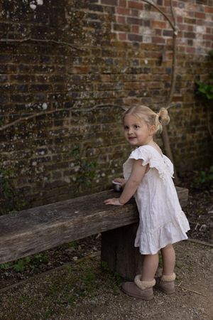 Toddler girl in white dress and brown boots standing by a rustic wooden bench against an old brick wall.