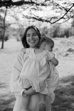Black and white photo of a smiling woman in a white dress holding a young child outdoors near trees, photographed in Brighton