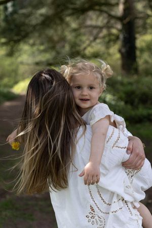 A mother in a white dress holds her smiling toddler girl outdoors in a lush green forest setting during a Brighton family photoshoot