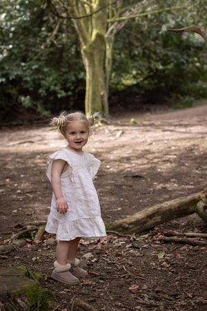 Toddler girl in white dress and tan boots smiling in woodland setting during a Brighton family photography session